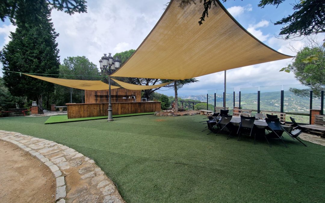 Área de bar al aire libre con toldos vela y césped artificial en la terraza de The Castle Barcelona, con vistas panorámicas a la montaña.